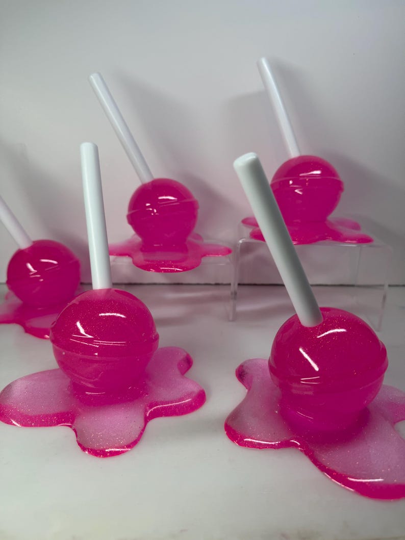 Puede incluir: Varias piruletas de color rosa brillante con palitos blancos. Cada piruleta se asienta sobre una base rosa con aspecto de derretida. Las piruletas tienen un aspecto brillante y las bases tienen un efecto brillante.