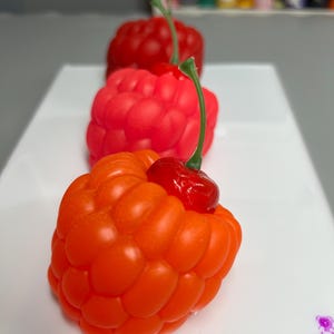 May include: Three colorful, berry-shaped objects in a row, each with a green stem. The front berry is orange, followed by a red one, and then a darker red one in the background. The objects are on a white surface.