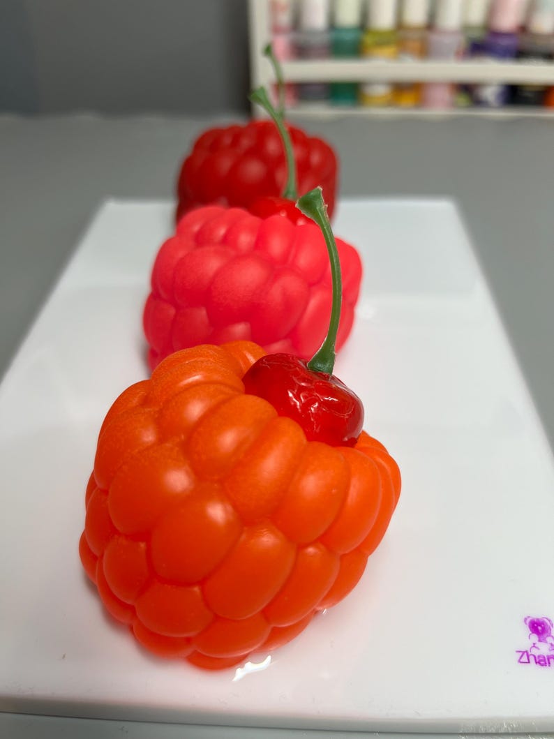May include: Three colorful, berry-shaped objects in a row, each with a green stem. The front berry is orange, followed by a red one, and then a darker red one in the background. The objects are on a white surface.