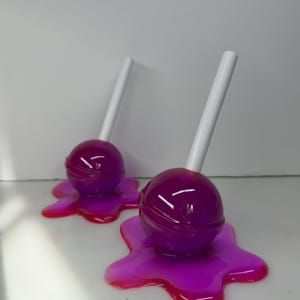 May include: Two purple lollipops with white sticks, resting on a vibrant, melted, pink and red base. The lollipops have a glossy finish, and the background is a plain white surface.
