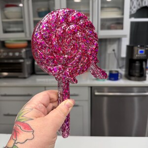 May include: A lollipop-shaped decorative object made of clear resin filled with vibrant pink and purple glitter. The circular head features a swirling pattern with a small protruding piece on the right side, attached to a straight, rectangular handle.