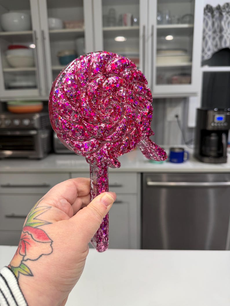 May include: A lollipop-shaped decorative object made of clear resin filled with vibrant pink and purple glitter. The circular head features a swirling pattern with a small protruding piece on the right side, attached to a straight, rectangular handle.