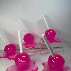 Puede incluir: Varias piruletas rosas con palitos blancos, cada una sobre una base rosa a juego, brillante y con aspecto derretido. Las piruletas est&aacute;n dispuestas sobre una superficie blanca, algunas elevadas sobre soportes transparentes.