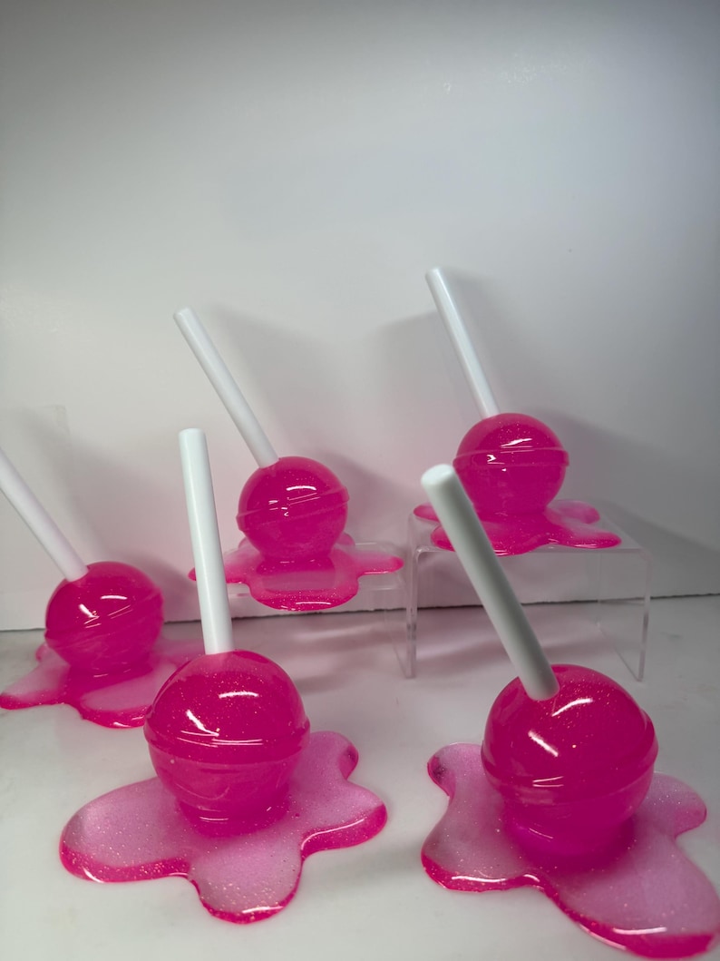 Puede incluir: Varias piruletas rosas con palitos blancos, cada una sobre una base rosa a juego, brillante y con aspecto derretido. Las piruletas est&aacute;n dispuestas sobre una superficie blanca, algunas elevadas sobre soportes transparentes.