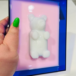 Puede incluir: Una pieza de arte mural rectangular que presenta una figura tridimensional de oso de goma blanco brillante centrada sobre un fondo rosa claro liso, todo ello encerrado en un marco de acr&iacute;lico azul oscuro transl&uacute;cido con bordes definidos.