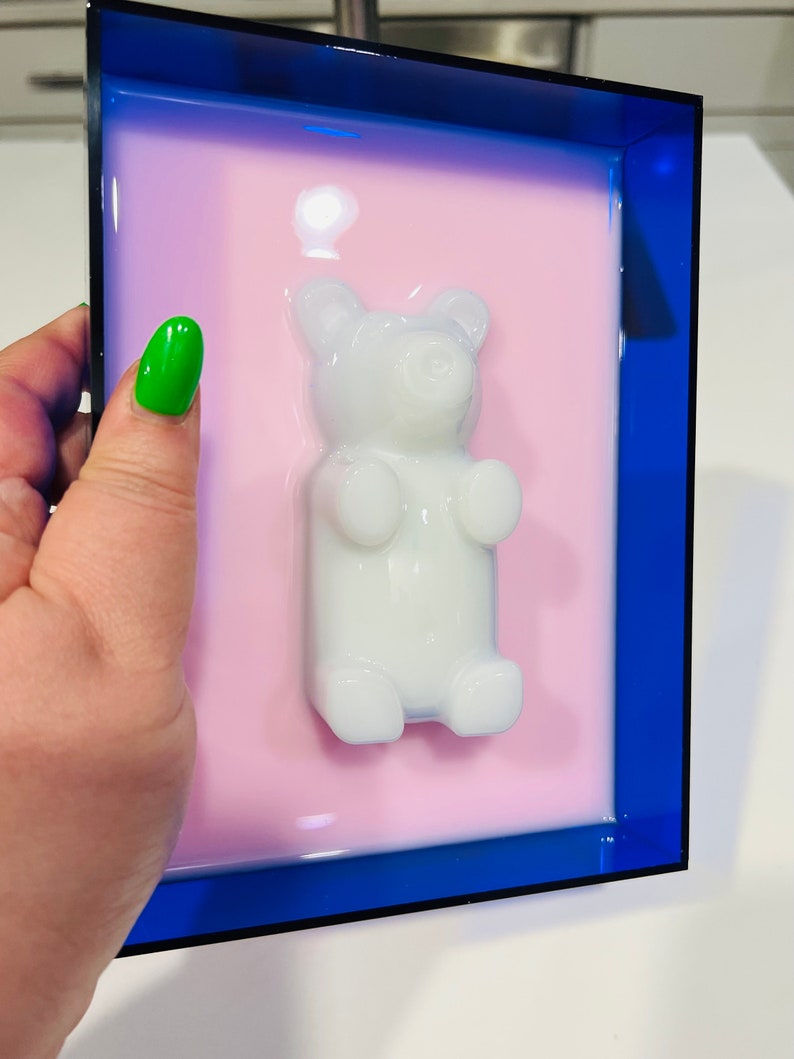 Puede incluir: Una pieza de arte mural rectangular que presenta una figura tridimensional de oso de goma blanco brillante centrada sobre un fondo rosa claro liso, todo ello encerrado en un marco de acr&iacute;lico azul oscuro transl&uacute;cido con bordes definidos.