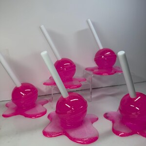 Puede incluir: Una colecci&oacute;n de piruletas rosas con palitos blancos. Cada piruleta tiene una parte superior redonda y brillante y se asienta sobre una base rosa y brillante que se asemeja a una forma derretida. Las piruletas est&aacute;n dispuestas sobre una superficie blanca.