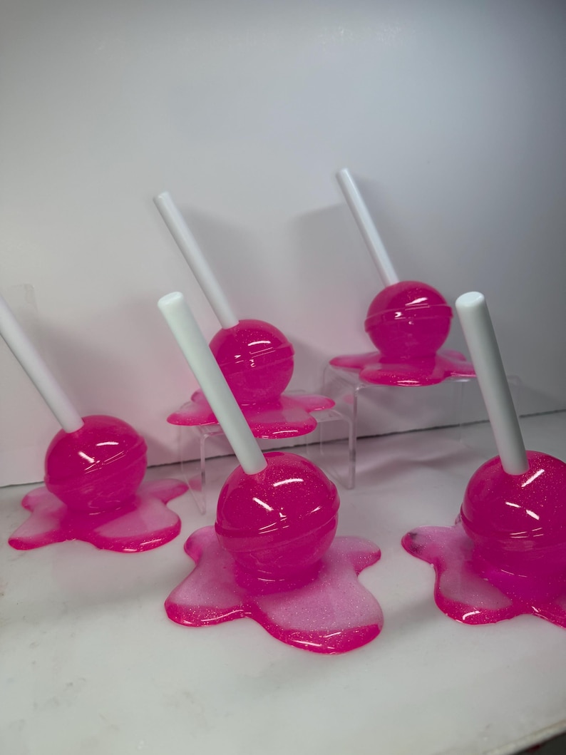 Puede incluir: Una colecci&oacute;n de piruletas rosas con palitos blancos. Cada piruleta tiene una parte superior redonda y brillante y se asienta sobre una base rosa y brillante que se asemeja a una forma derretida. Las piruletas est&aacute;n dispuestas sobre una superficie blanca.