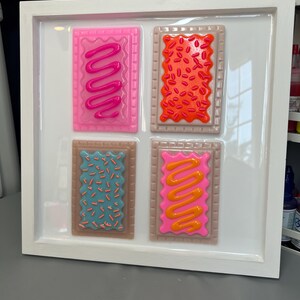 May include: A framed artwork featuring four colorful, stylized Pop-Tart designs. Each rectangular pastry has a different frosting and sprinkle pattern in shades of pink, orange, blue, and brown. The artwork is set against a white background within a white frame.