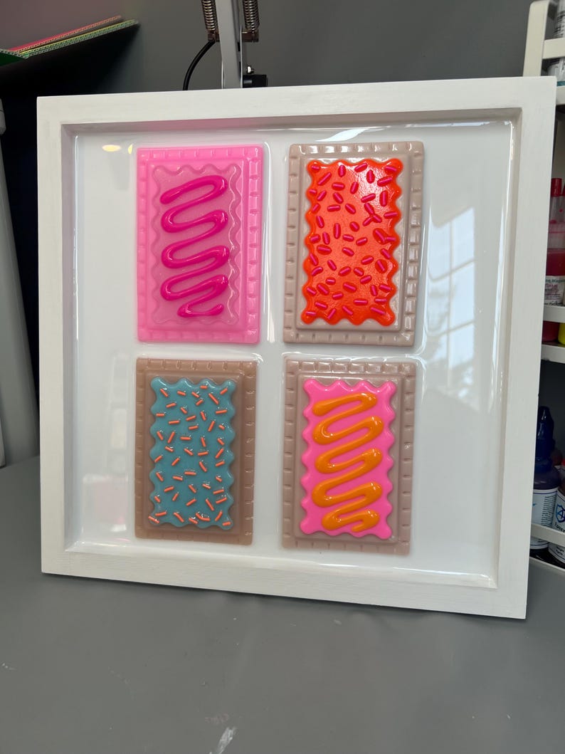 May include: A framed artwork featuring four colorful, stylized Pop-Tart designs. Each rectangular pastry has a different frosting and sprinkle pattern in shades of pink, orange, blue, and brown. The artwork is set against a white background within a white frame.