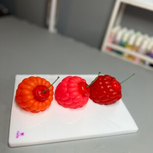 May include: Three colorful, fruit-shaped candles on a white rectangular plate. The candles are shaped like a raspberry, a cherry, and a raspberry-cherry hybrid. The raspberry is red, the cherry is red, and the hybrid is orange and red.