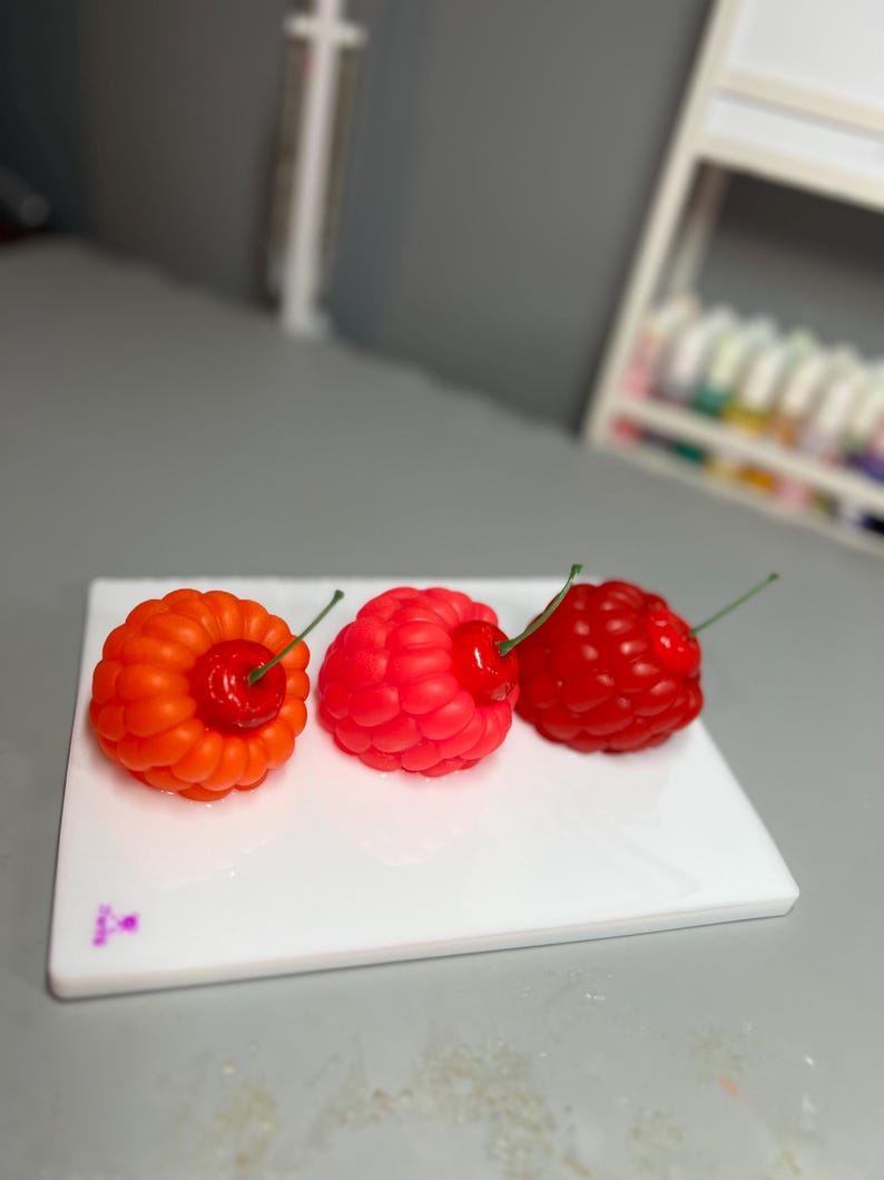 May include: Three colorful, fruit-shaped candles on a white rectangular plate. The candles are shaped like a raspberry, a cherry, and a raspberry-cherry hybrid. The raspberry is red, the cherry is red, and the hybrid is orange and red.