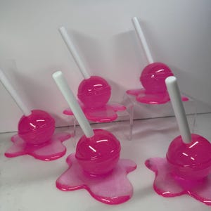 Puede incluir: Piruletas rosas con palitos blancos. Cada piruleta tiene una cabeza de caramelo rosa brillante y redonda, colocada sobre una base rosa a juego con forma de goteo. La imagen presenta un producto con tem&aacute;tica de caramelos, ideal para regalos.