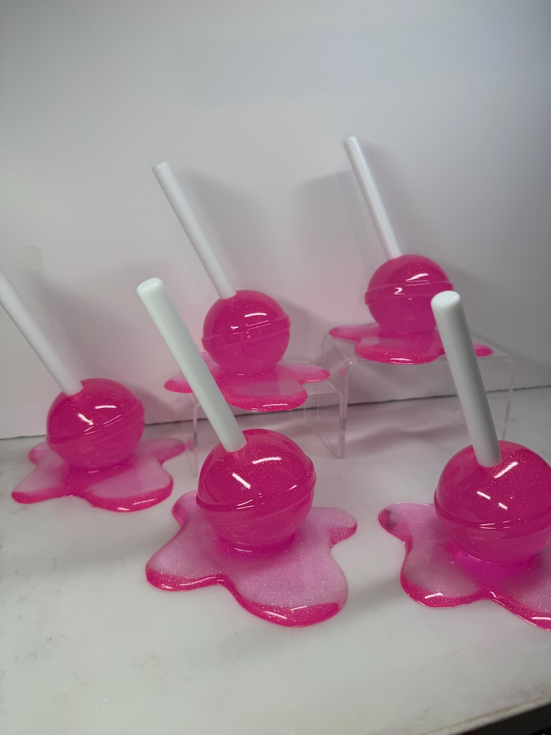 Puede incluir: Piruletas rosas con palitos blancos. Cada piruleta tiene una cabeza de caramelo rosa brillante y redonda, colocada sobre una base rosa a juego con forma de goteo. La imagen presenta un producto con tem&aacute;tica de caramelos, ideal para regalos.