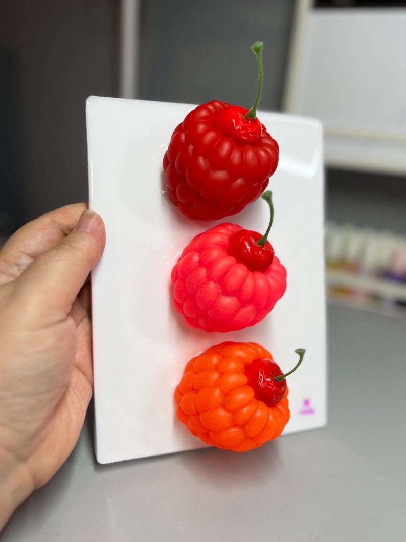 May include: Three colorful raspberry-shaped objects in red, pink, and orange are displayed on a white rectangular surface. Each raspberry has a small red cherry on top with a green stem. The objects are held by a hand.