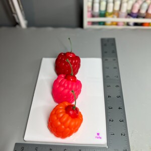 May include: Three colorful, raspberry-shaped candles on a white surface. The candles are red, pink, and orange, each with a green stem and a small red cherry on top. A metal ruler is placed next to the candles.