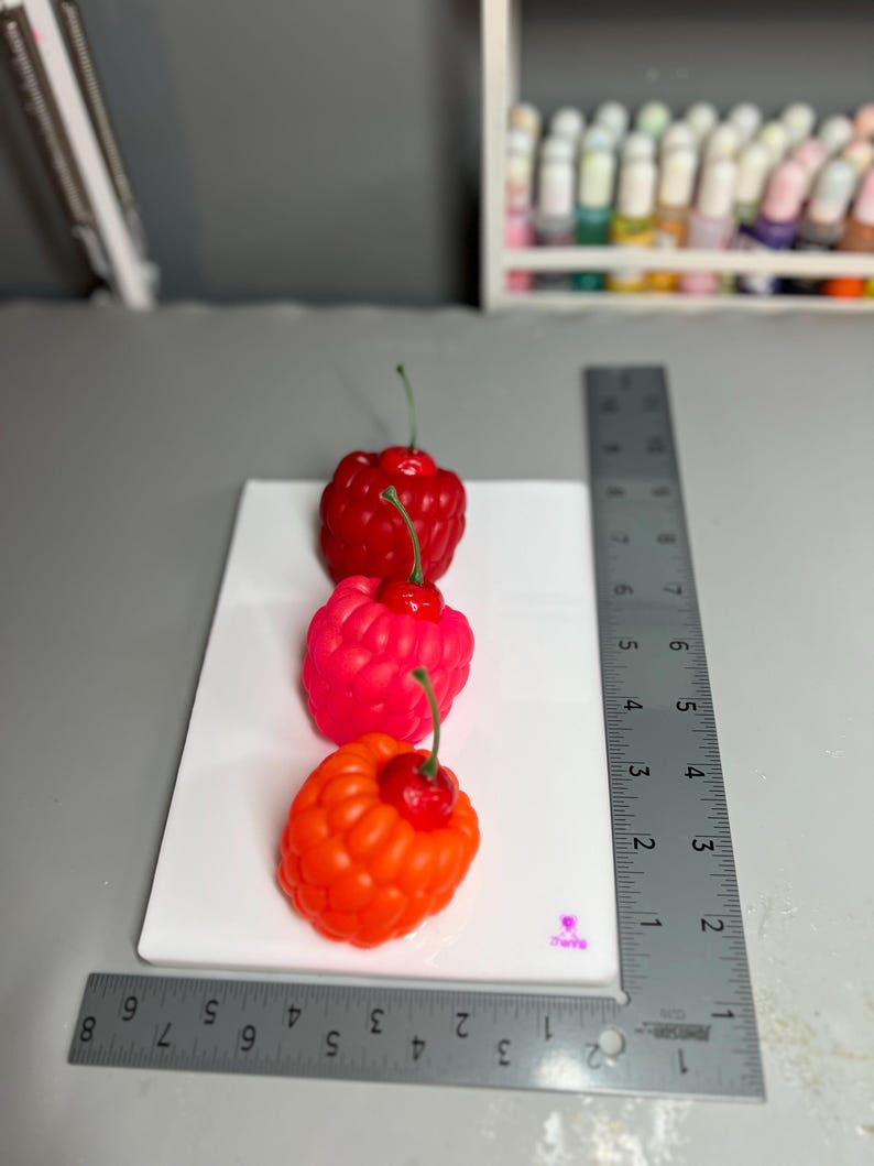 May include: Three colorful, raspberry-shaped candles on a white surface. The candles are red, pink, and orange, each with a green stem and a small red cherry on top. A metal ruler is placed next to the candles.
