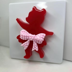 May include: A red bear-shaped decorative item with a pink bow, mounted on a white square. The bear is in a dancing pose, with arms outstretched. The bow has a textured, dotted pattern.