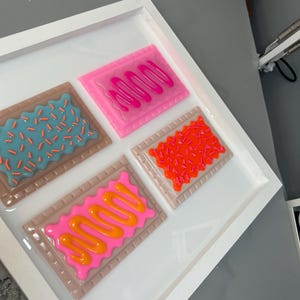 May include: A white framed artwork featuring four colorful, stylized Pop-Tart designs. Each pastry has a different frosting and sprinkle pattern, including pink, orange, and blue. The artwork is set against a white background.