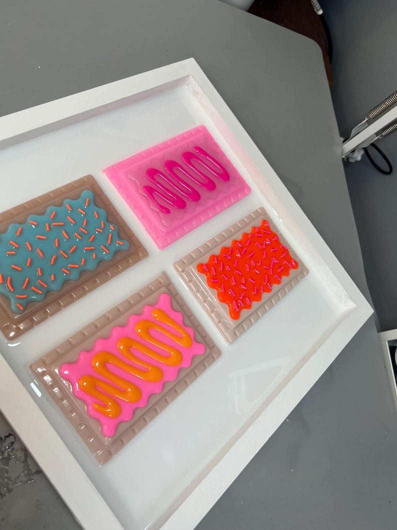 May include: A white framed artwork featuring four colorful, stylized Pop-Tart designs. Each pastry has a different frosting and sprinkle pattern, including pink, orange, and blue. The artwork is set against a white background.