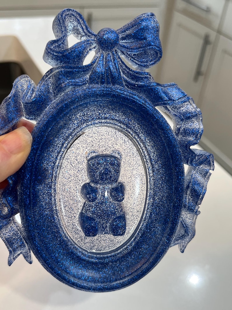 Op de afbeelding: Een blauw glitterhars fotolijst met een witte binnenkant en een blauw glitterbeer silhouet erin. De lijst is versierd met een blauw glitter strikje.