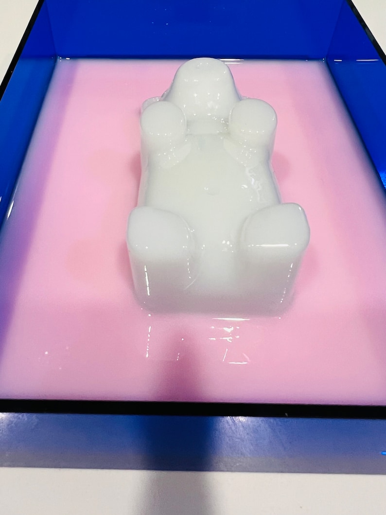 Puede incluir: Un objeto blanco, semitransl&uacute;cido, con forma de oso, que descansa en un l&iacute;quido rosa claro. El oso est&aacute; colocado dentro de un recipiente rectangular con un borde azul oscuro. La superficie del oso es lisa, redondeada y refleja la luz.