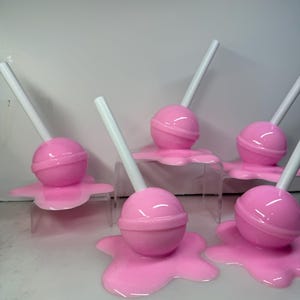 Op de afbeelding: Een verzameling roze lolly's met witte stokjes, die lijken te smelten op een helder oppervlak. De lolly's hebben een glanzende afwerking en zijn in een groep gerangschikt, wat een speelse en grillige esthetiek cre&euml;ert.