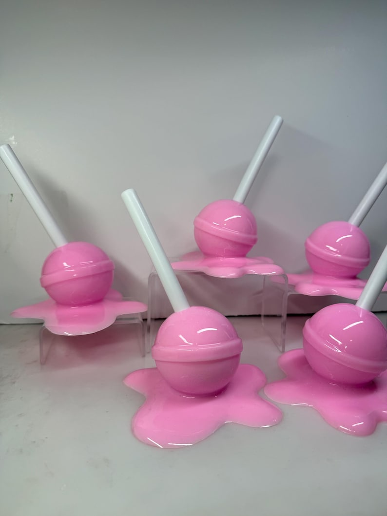 Op de afbeelding: Een verzameling roze lolly's met witte stokjes, die lijken te smelten op een helder oppervlak. De lolly's hebben een glanzende afwerking en zijn in een groep gerangschikt, wat een speelse en grillige esthetiek cre&euml;ert.
