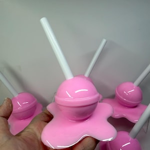 Op de afbeelding: Verschillende roze objecten in de vorm van lolly's met witte stokjes. De lolly's hebben een glanzende afwerking en staan op een gesmolten uitziende basis. De afbeelding is een close-up, die de details van het snoepgoed laat zien.