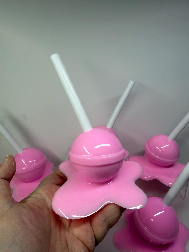 Op de afbeelding: Verschillende roze objecten in de vorm van lolly's met witte stokjes. De lolly's hebben een glanzende afwerking en staan op een gesmolten uitziende basis. De afbeelding is een close-up, die de details van het snoepgoed laat zien.