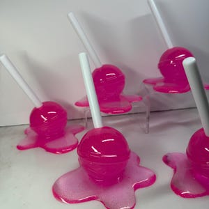 Puede incluir: Varios caramelos rosas con palitos blancos. Cada caramelo est&aacute; sobre una base rosa brillante y derretida. Los caramelos est&aacute;n colocados sobre una superficie blanca, algunos sobre un soporte acr&iacute;lico transparente.