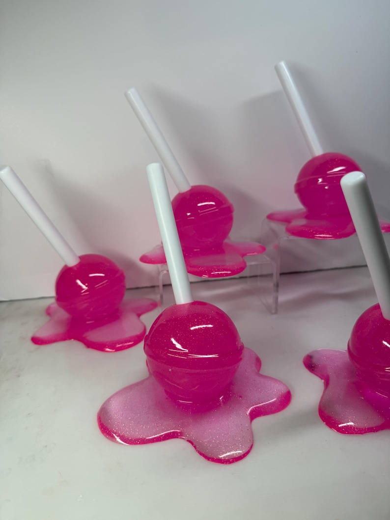 Puede incluir: Varios caramelos rosas con palitos blancos. Cada caramelo est&aacute; sobre una base rosa brillante y derretida. Los caramelos est&aacute;n colocados sobre una superficie blanca, algunos sobre un soporte acr&iacute;lico transparente.