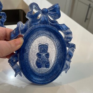 Op de afbeelding: Een blauwe en witte glitter fotolijst met een decoratieve strik en een wit silhouet van een teddybeer erin.