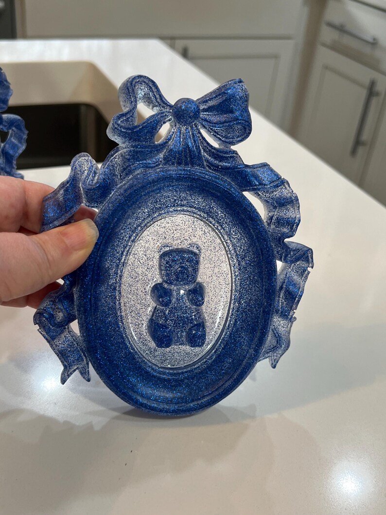 Op de afbeelding: Een blauwe en witte glitter fotolijst met een decoratieve strik en een wit silhouet van een teddybeer erin.