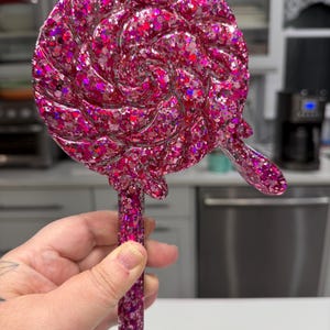 May include: A circular lollipop-shaped object featuring a swirling pattern and a thick handle, entirely covered in a dense layer of reflective pink and magenta glitter. The surface has a glossy, smooth finish with a small drip detail on the side.