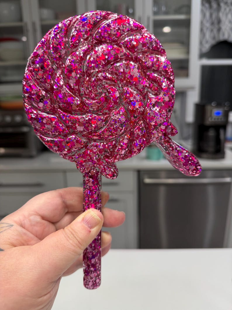 May include: A circular lollipop-shaped object featuring a swirling pattern and a thick handle, entirely covered in a dense layer of reflective pink and magenta glitter. The surface has a glossy, smooth finish with a small drip detail on the side.