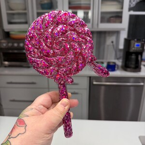 May include: A handheld mirror shaped like a round lollipop with a swirl design. The mirror is made of a translucent pink resin material embedded with numerous reflective pink and silver glitter flakes throughout the entire surface and handle.