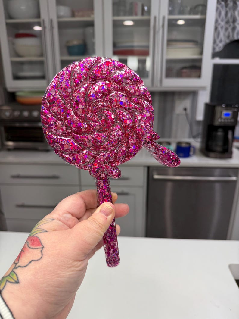 May include: A handheld mirror shaped like a round lollipop with a swirl design. The mirror is made of a translucent pink resin material embedded with numerous reflective pink and silver glitter flakes throughout the entire surface and handle.