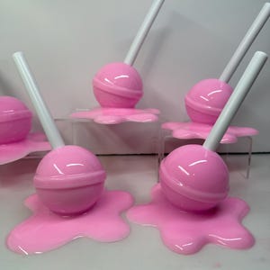 Op de afbeelding: Roze lolly's met witte stokjes, die lijken te smelten op een bijpassende roze ondergrond. De lolly's zijn geplaatst op transparante acrylstandaards, wat een speelse uitstraling cre&euml;ert. De snoep-thema objecten staan tegen een witte achtergrond.