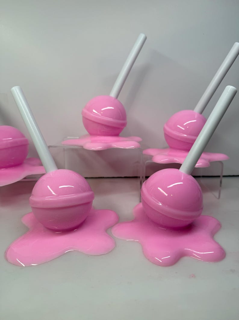 Op de afbeelding: Roze lolly's met witte stokjes, die lijken te smelten op een bijpassende roze ondergrond. De lolly's zijn geplaatst op transparante acrylstandaards, wat een speelse uitstraling cre&euml;ert. De snoep-thema objecten staan tegen een witte achtergrond.