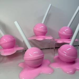 Op de afbeelding: Roze lolly-vormige decoraties met witte stokjes, rustend op een bijpassende roze, gesmolten uitziende basis. De lolly's zijn gerangschikt op heldere, verhoogde platforms, wat een speelse en grillige esthetiek cre&euml;ert.