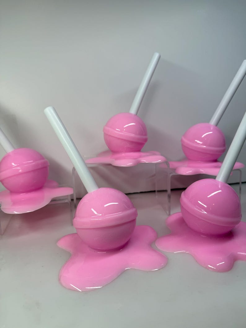 Op de afbeelding: Roze lolly-vormige decoraties met witte stokjes, rustend op een bijpassende roze, gesmolten uitziende basis. De lolly's zijn gerangschikt op heldere, verhoogde platforms, wat een speelse en grillige esthetiek cre&euml;ert.