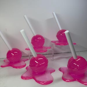 Puede incluir: Una colecci&oacute;n de piruletas rosas con palitos blancos, cada una de las cuales parece estar derriti&eacute;ndose sobre una base rosa a juego. Las piruletas tienen un acabado brillante y las bases tienen un efecto brillante. El fondo es blanco liso.