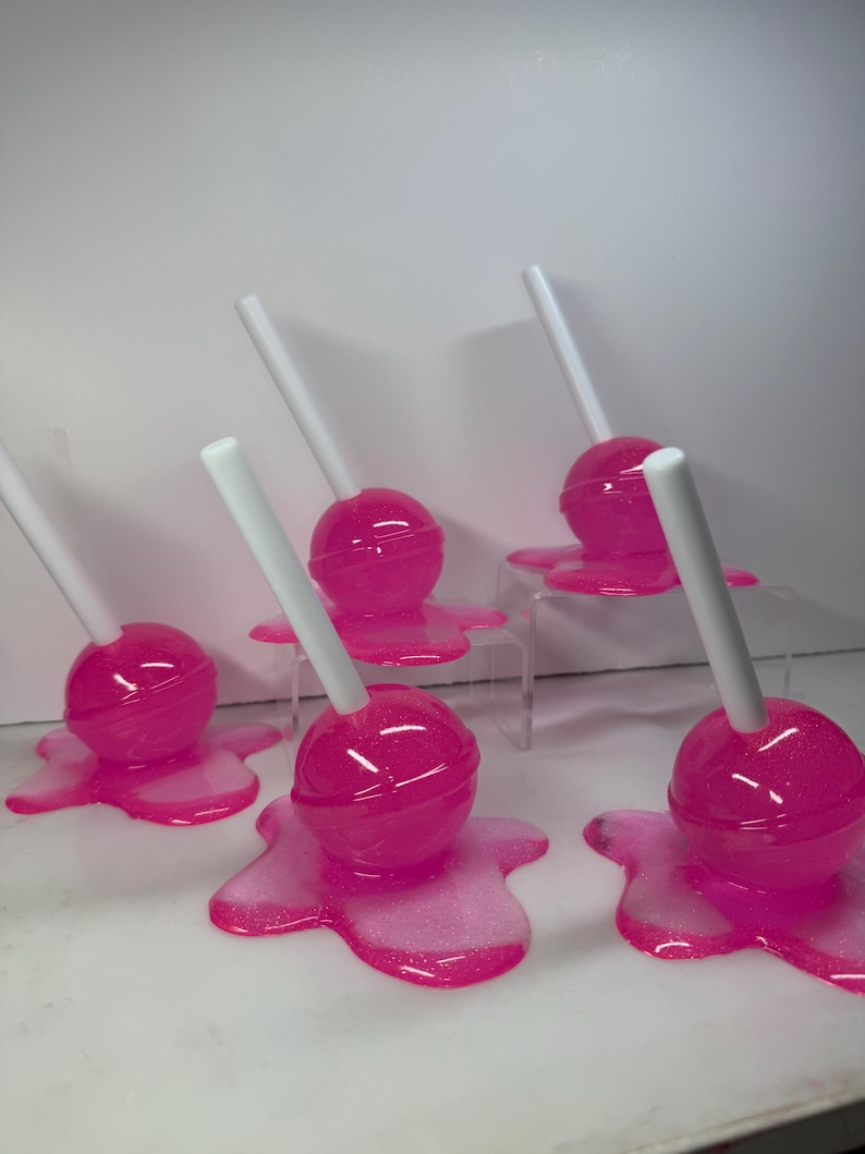 Puede incluir: Una colecci&oacute;n de piruletas rosas con palitos blancos, cada una de las cuales parece estar derriti&eacute;ndose sobre una base rosa a juego. Las piruletas tienen un acabado brillante y las bases tienen un efecto brillante. El fondo es blanco liso.