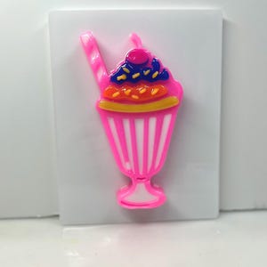 May include: A pink sundae-shaped decoration with a cherry on top. The sundae has a pink and white striped cup, with layers of orange, blue, and yellow toppings. A pink straw extends from the sundae.