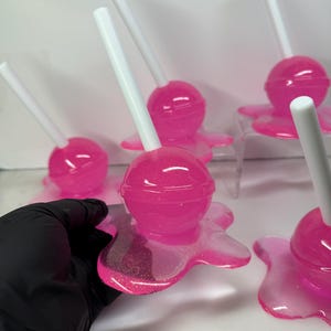 Puede incluir: Varias piezas de arte de resina rosa con forma de piruleta, con palitos blancos. Cada piruleta se asienta sobre una base rosa con aspecto de derretida. La resina tiene un acabado brillante, sobre un fondo blanco.