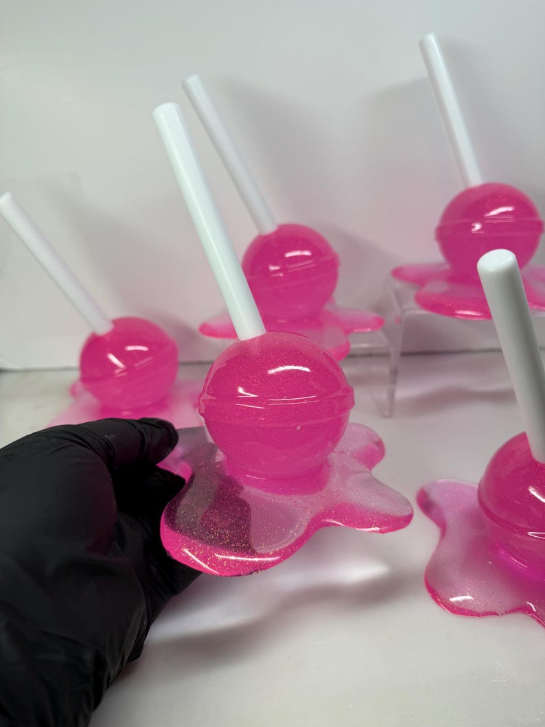Puede incluir: Varias piezas de arte de resina rosa con forma de piruleta, con palitos blancos. Cada piruleta se asienta sobre una base rosa con aspecto de derretida. La resina tiene un acabado brillante, sobre un fondo blanco.