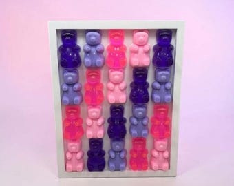 May include: A white rectangular shadow box frame containing a grid of twenty translucent gummy bear figures arranged in five rows and four columns. The bears are colored in various shades of pink, purple, blue, and magenta, creating a vibrant display.