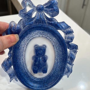 Op de afbeelding: Een blauw glitterhars fotolijst met een witte ovale passe-partout en een blauwe gummybeer erin. De lijst is versierd met een blauw glitter strikje.