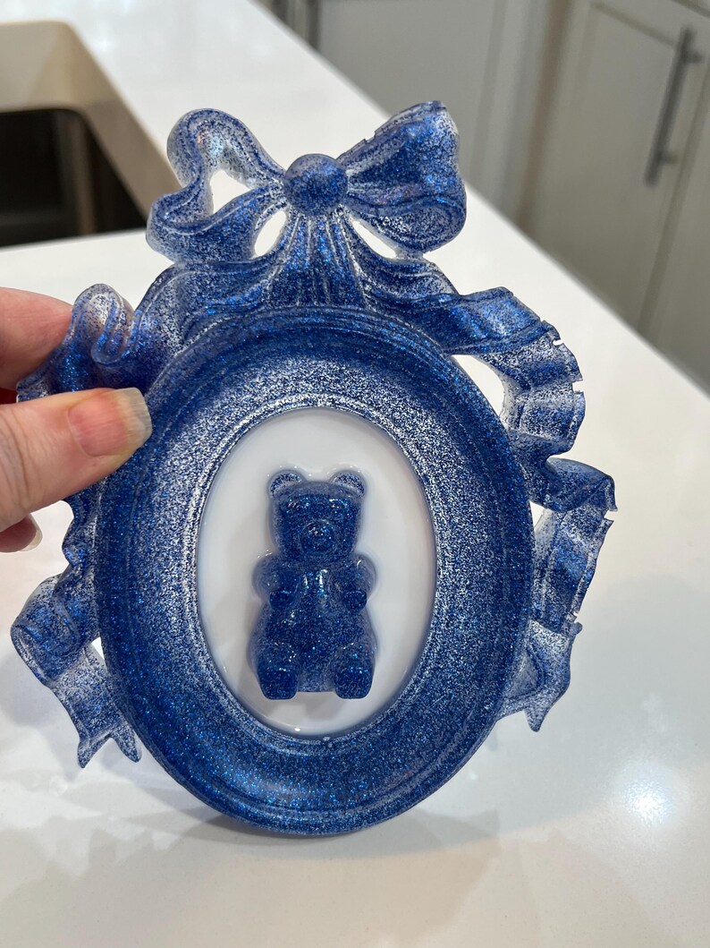 Op de afbeelding: Een blauw glitterhars fotolijst met een witte ovale passe-partout en een blauwe gummybeer erin. De lijst is versierd met een blauw glitter strikje.